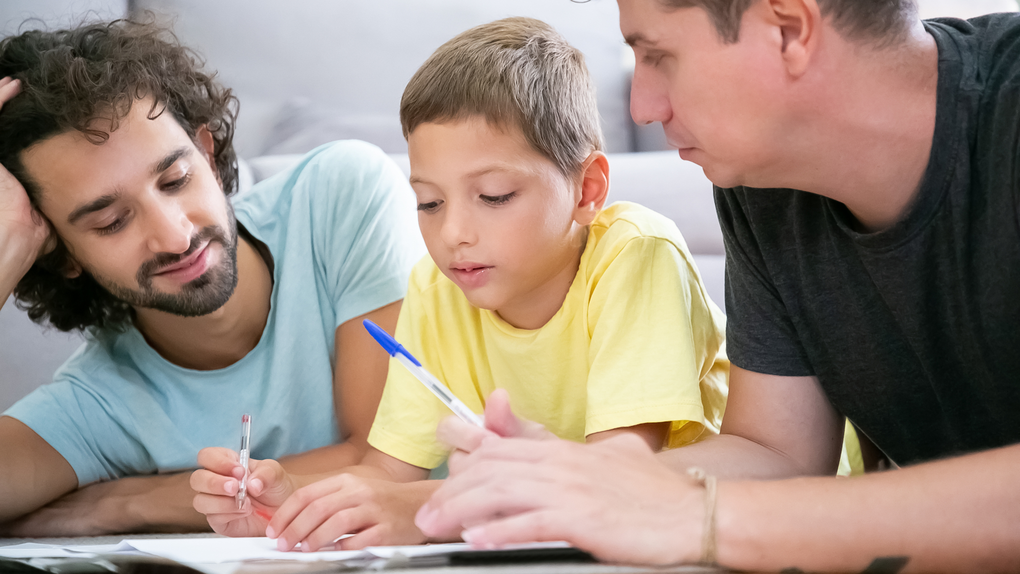 Two fathers helping focused boy with school home task, lying on floor at home, writing or drawing in papers. Family and gay parents concept